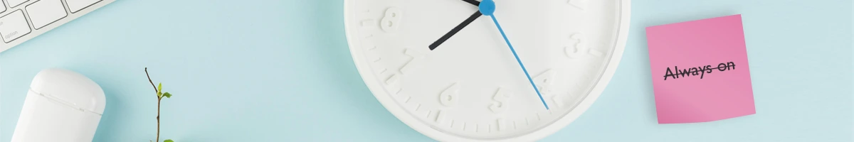 Set boundaries in email signatures A birds-eye-view of a desk with various stationary and a clock with a 'Always on' post-it that's been scratched out.